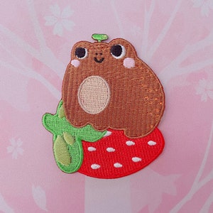 May include: A brown frog with a green leaf on its head sits on a red strawberry with white seeds. The frog has a happy expression and pink cheeks.