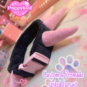 Può includere: Un collare in finta pelliccia nera con punte rosa e una fibbia in plastica rosa. Il collare è progettato per fursuit e presenta il testo "Custom & Premade spiked fursuit collars". L'immagine ha un'estetica rosa e bianca.