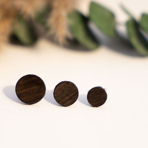 Può includere: Sono presentati tre orecchini a bottone in legno marrone scuro di diverse dimensioni. Gli orecchini circolari mostrano un motivo a venature del legno. Lo sfondo è una superficie bianca con vegetazione sfocata e piante secche.