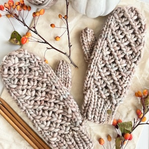 Könnte beinhalten: Ein Paar grob gestrickte Fäustlinge in hellen Beige- und Grautönen. Die Fäustlinge werden mit Stricknadeln aus Holz, einem Wollknäuel und einer weißen Tasse präsentiert. Herbstliche Beeren und Zweige ergänzen die Szene.