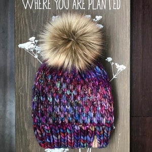 May include: A colourful knitted beanie with a fluffy brown pom pom. The beanie features a variety of colours, including purple, blue, pink, and green. The beanie is displayed on a wooden surface with a sign that reads "Bloom where you are planted".
