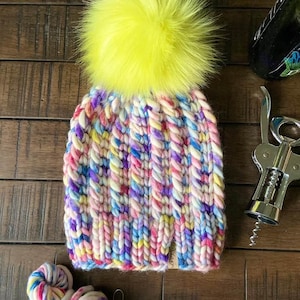 May include: A colorful knitted beanie with a bright yellow faux fur pom pom. The beanie is made with a variety of colors, including pink, blue, purple, and yellow. The beanie is sitting on a wooden surface.