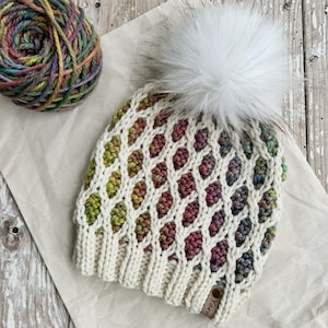 May include: Cream-colored knit hat with a diamond pattern and a large white faux fur pom-pom. The hat features a ribbed brim and is adorned with colorful yarn accents. A ball of multi-colored yarn sits beside the hat.