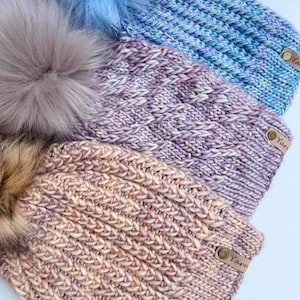 May include: Three knitted hats with faux fur pom poms. The hats are in shades of pink, purple, and blue. Each hat has a leather tag with the word "Yarn" on it.