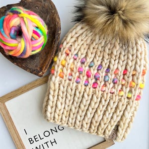 May include: A cream-colored knit hat with colorful pom-poms and a large faux fur pom-pom on top. A wooden heart-shaped bowl holds a skein of rainbow yarn. A framed sign reads "I BELONG WITH YOU."