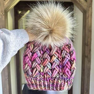 May include: A hand-knitted beanie in a vibrant mix of pink, purple, and yellow yarns, topped with a large, fluffy, tan-coloured pom-pom. The hat features a textured, woven design and a small tag.