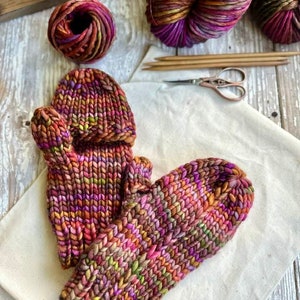 Könnte beinhalten: Ein Paar gestrickte Fingerlose Handschuhe in einem mehrfarbigen Garn mit Schattierungen von Lila, Rosa, Braun und Grün. Das Garn ist ein dickes Gewicht und die Handschuhe sind mit einem einfachen Stichmuster gefertigt.