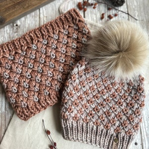 May include: A knitted hat and matching scarf in a brown and beige color scheme. The hat has a fluffy pompom and a leather tag that says "Yarn".