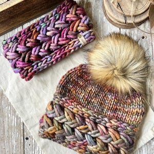 May include: Two colourful knitted hats with a braided pattern. One hat is a headband style and the other is a beanie with a faux fur pom pom. Both hats have a brown leather tag with the word "Loopy" on it.