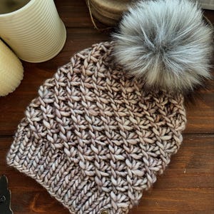 Peut inclure: Un bonnet tricoté à la main dans des tons de marron et de crème, surmonté d'un gros pompon en fausse fourrure grise. Le bonnet présente un motif de point texturé et une petite étiquette ronde avec du texte.
