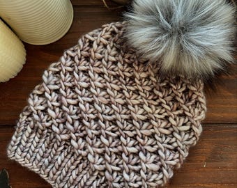 May include: A hand-knitted beanie in shades of brown and cream, with a large, fluffy grey faux fur pom-pom. The hat has a textured stitch pattern and a small, round tag with text. The beanie is on a wooden surface.