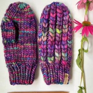 Puede incluir: Un par de mitones de punto coloridos con un esquema de color morado, rosa y azul. Los mitones tienen un patrón de punto texturizado y están hechos con un hilo grueso. Los mitones están sobre una superficie blanca con agujas de tejer de madera y una flor.