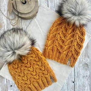 Puede incluir: Dos gorros de punto color amarillo mostaza con un patrón de chevrón texturizado, un borde acanalado y un gran pompón de piel sintética esponjoso en tonos gris y blanco. Cada gorro incluye una pequeña etiqueta rectangular con la palabra Yarn.
