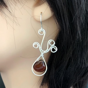 May include: Silver wire earrings with a unique design. Each earring features a teardrop-shaped, reddish-brown gemstone suspended within a wire frame. The wirework includes decorative swirls and a hook for pierced ears.