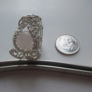 May include: A handcrafted silver wire-wrapped ring with a light pink rose quartz stone. The ring is next to a dime for size comparison. The ring's design features intricate weaving and a delicate, feminine aesthetic.