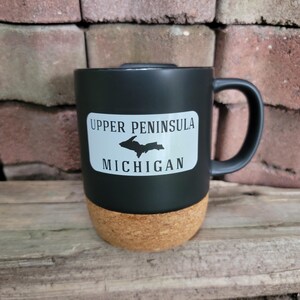 May include: Black travel mug with a cork bottom and a white sticker that says "Upper Peninsula Michigan" with an outline of the Upper Peninsula of Michigan.