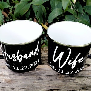May include: Two black enamel mugs with white lettering. The mug on the left says "Husband Est. 11.27.2021" and the mug on the right says "Wife Est. 11.27.2021".