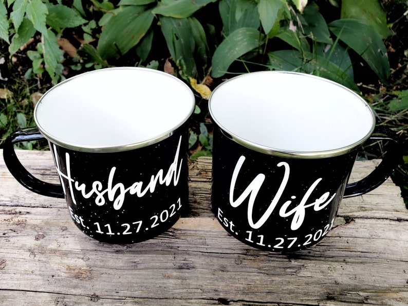 May include: Two black enamel mugs with white lettering. The mug on the left says "Husband Est. 11.27.2021" and the mug on the right says "Wife Est. 11.27.2021".