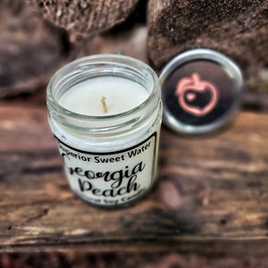 May include: A clear glass jar candle with a white wax fill and a single wick. The label reads Superior Sweet Water Georgia Peach Natural Soy Candle. A separate silver metal lid sits nearby, featuring a peach icon with a small heart in the center.