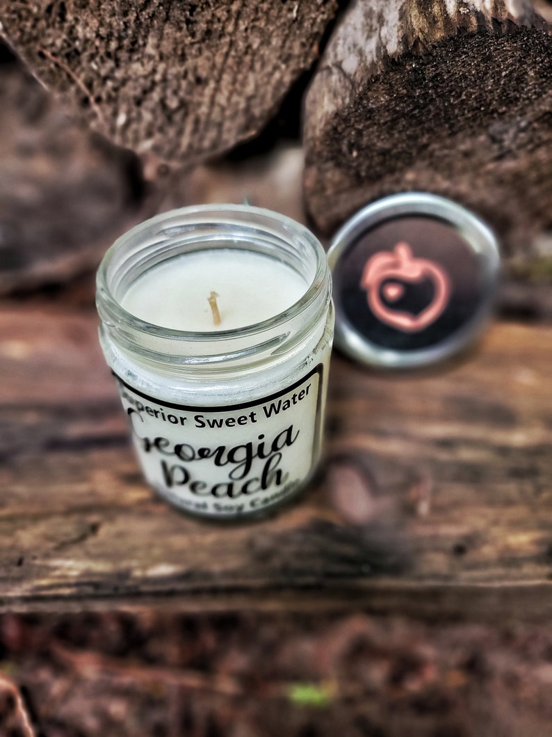 May include: A clear glass jar candle with a white wax fill and a single wick. The label reads Superior Sweet Water Georgia Peach Natural Soy Candle. A separate silver metal lid sits nearby, featuring a peach icon with a small heart in the center.