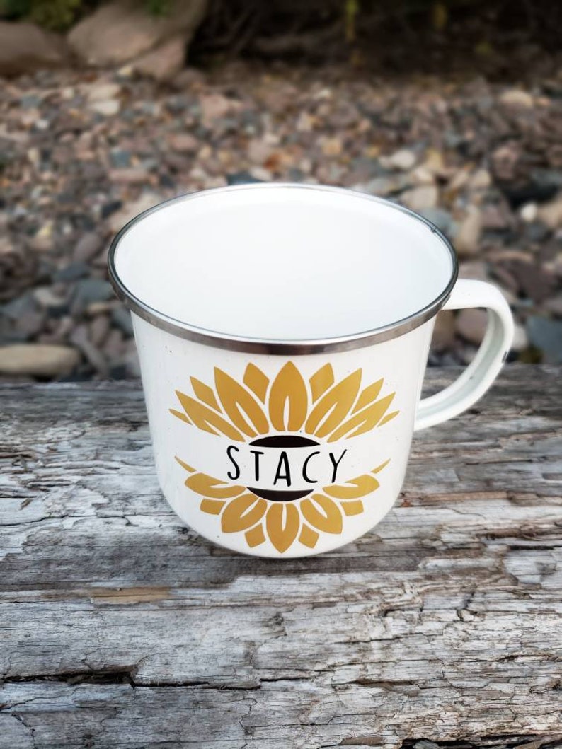 Op de afbeelding: Een witte emaille mok met een gouden zonnebloemontwerp en de naam "Stacy" in zwart op een witte banner.