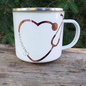 May include: White enamel mug with a copper-colored heart and stethoscope design. The word "Savannah" is written inside the heart.