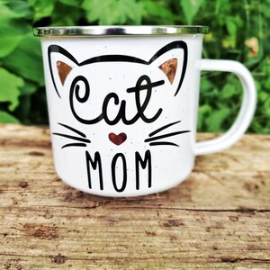 May include: White enamel mug with a black and white cat design. The cat has a red heart nose and the text "Cat Mom" is printed below the cat.