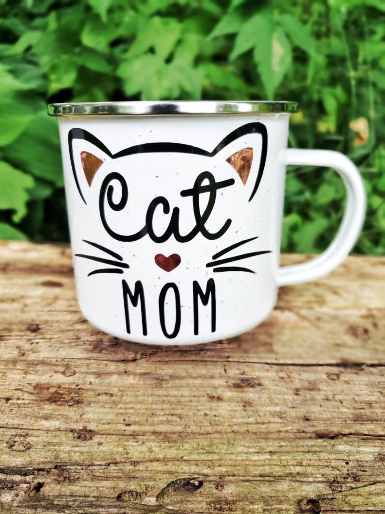May include: White enamel mug with a black and white cat design. The cat has a red heart nose and the text "Cat Mom" is printed below the cat.