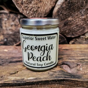 May include: A clear glass jar candle with a silver metal lid containing white wax. The label features black text reading Superior Sweet Water, Georgia Peach, and Natural Soy Candle, framed by a thin black border on a cream-colored background.