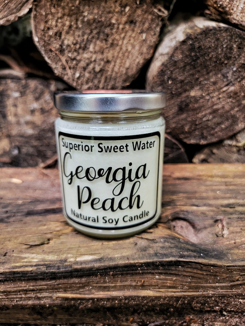 May include: A clear glass jar candle with a silver metal lid containing white wax. The label features black text reading Superior Sweet Water, Georgia Peach, and Natural Soy Candle, framed by a thin black border on a cream-colored background.