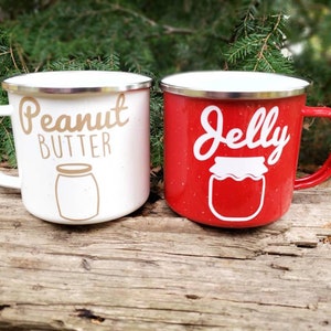May include: Two enamel camping mugs with a white and red speckled finish. The white mug has the text "Peanut Butter" in gold lettering with a white outline and a brown jar illustration. The red mug has the text "Jelly" in white lettering with a white outline and a white jar illustration.