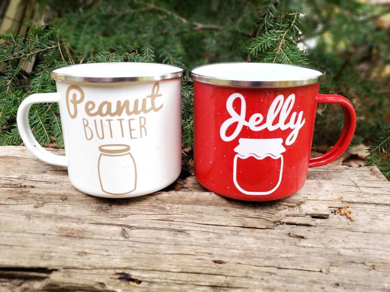 May include: Two enamel camping mugs with a white and red speckled finish. The white mug has the text "Peanut Butter" in gold lettering with a white outline and a brown jar illustration. The red mug has the text "Jelly" in white lettering with a white outline and a white jar illustration.