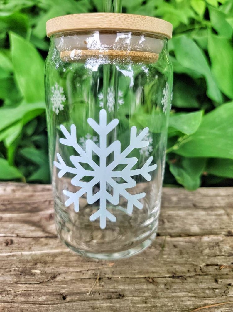 May include: A clear glass drinking bottle with a bamboo lid and a white snowflake design. The bottle has a straw sticking out of the lid.