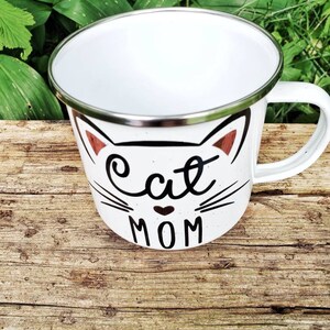 May include: White enamel mug with a black cat face and the words "Cat Mom" in black lettering. The mug has a silver rim.