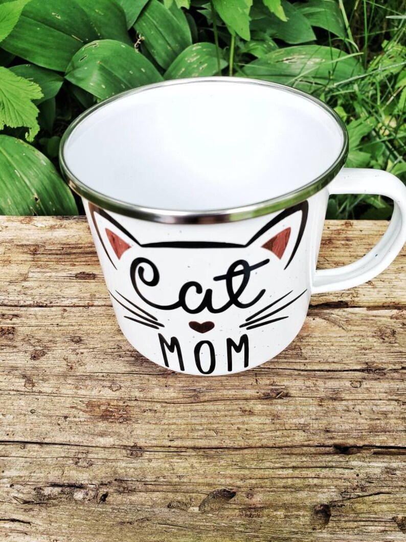 May include: White enamel mug with a black cat face and the words "Cat Mom" in black lettering. The mug has a silver rim.