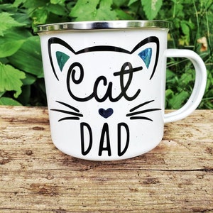May include: White enamel mug with a black cat face and the words "Cat Dad" in black lettering. The cat face has blue ears and a small black heart.