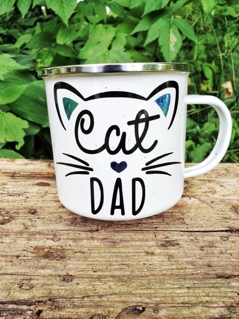 May include: White enamel mug with a black cat face and the words "Cat Dad" in black lettering. The cat face has blue ears and a small black heart.