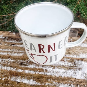 May include: White enamel camping mug with a silver rim. The mug has a red and silver design that says "Earned It".