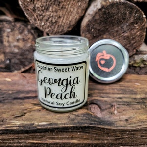 May include: A clear glass jar candle with a white label reading Superior Sweet Water Georgia Peach Natural Soy Candle. The silver lid features a peach icon with a small heart in the center. The candle is filled with white wax and has a single wick.