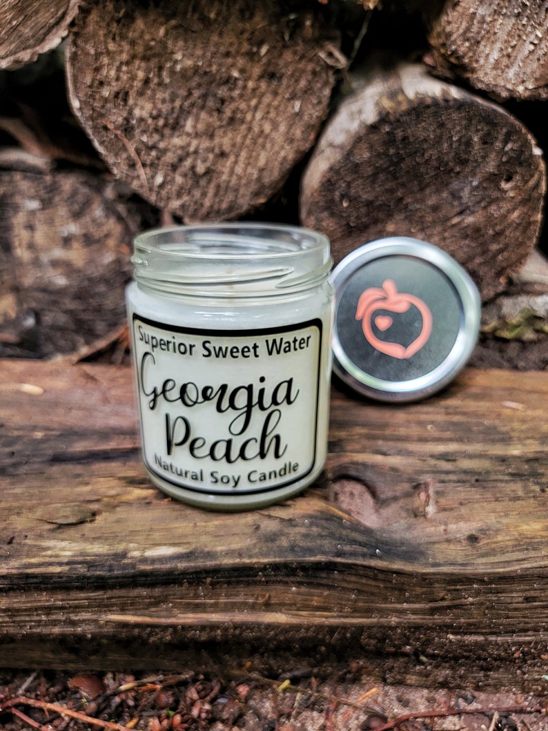 May include: A clear glass jar candle with a white label reading Superior Sweet Water Georgia Peach Natural Soy Candle. The silver lid features a peach icon with a small heart in the center. The candle is filled with white wax and has a single wick.