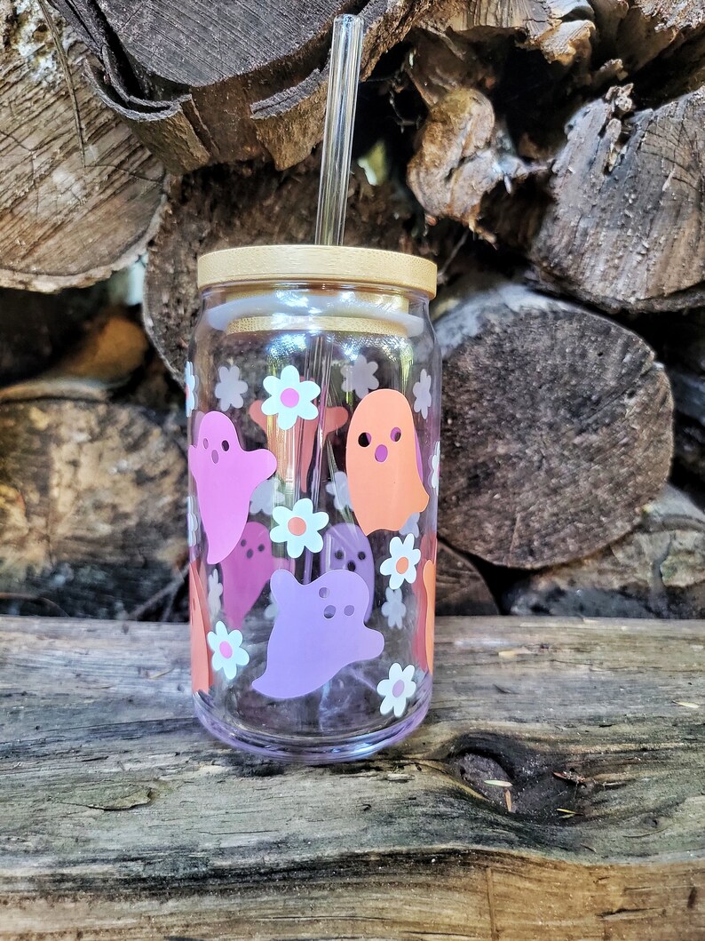 May include: A clear glass reusable water bottle with a bamboo lid and a straw. The bottle is decorated with pink, orange, and white ghost and flower designs.