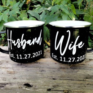 May include: Two black enamel mugs with white lettering. The mug on the left says "Husband Est. 11.27.2021" and the mug on the right says "Wife Est. 11.27.2021".