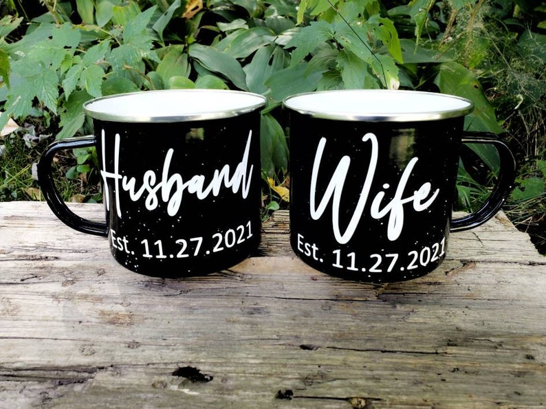 May include: Two black enamel mugs with white lettering. The mug on the left says "Husband Est. 11.27.2021" and the mug on the right says "Wife Est. 11.27.2021".