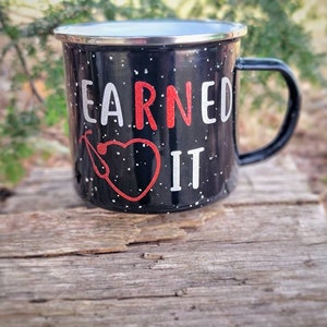 May include: Black enamel mug with white speckles and a red heart with a stethoscope. The mug says "EARNED IT" in white letters.