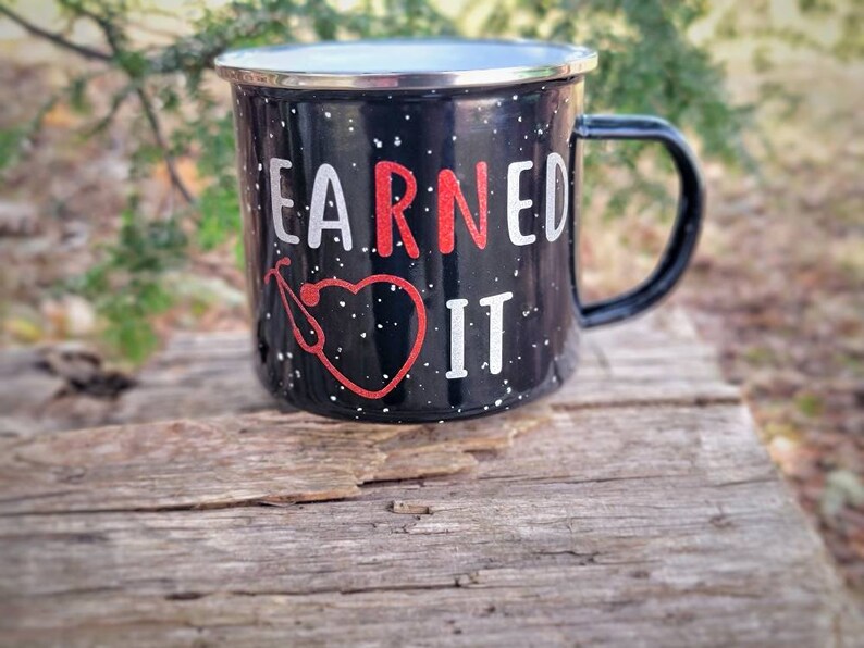May include: Black enamel mug with white speckles and a red heart with a stethoscope. The mug says "EARNED IT" in white letters.