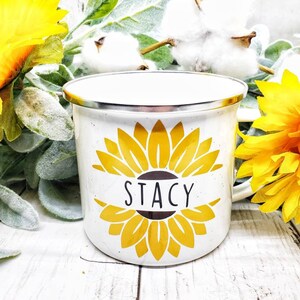 Op de afbeelding: Witte emaille mok met een geel zonnebloemontwerp en de naam "Stacy" in zwarte letters.