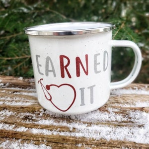 May include: White enamel mug with a red heart and stethoscope design. The text "EARNED IT" is printed on the mug in silver and red.