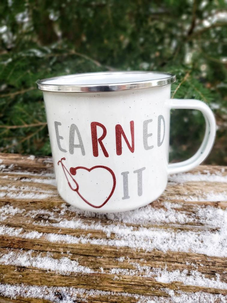 May include: White enamel mug with a red heart and stethoscope design. The text "EARNED IT" is printed on the mug in silver and red.