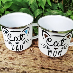 May include: Two white enamel mugs with black cat faces and the words "Cat Dad" and "Cat Mom" on them. The mugs are sitting on a wooden surface.