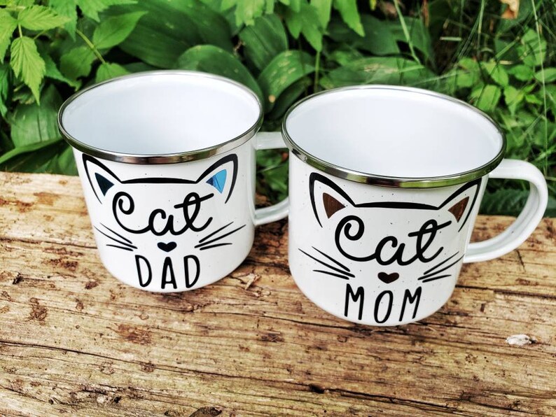 May include: Two white enamel mugs with black cat faces and the words "Cat Dad" and "Cat Mom" on them. The mugs are sitting on a wooden surface.